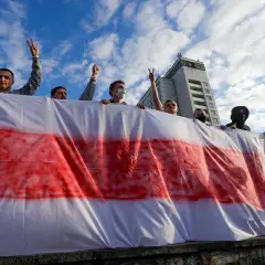 Ludzie trzymają biało-czerwoną flagę, symbol wolności i nadziei. Na niebie chmury, w tle budynek.