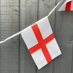 Girlanda z flagami Anglii wisi na szarej, drewnianej desce. Białe tło z czerwonym krzyżem.