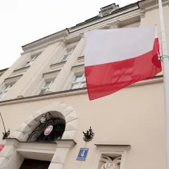Flaga Polski pionowo powiewa przed budynkiem. Biało-czerwone barwy dumne na tle jasnej fasady.