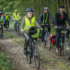Grupa rowerzystów w kamizelkach odblaskowych jedzie leśną ścieżką. Jedna z pań ma na rowerze zieloną flagę.