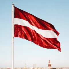 Flaga Łotwy: ciemnoczerwona z białą wstęgą pośrodku, na tle błękitnego nieba i miasta.