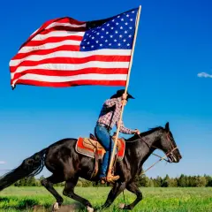 Jeździec w kapeluszu galopuje na czarnym koniu, niosąc pierwszą flagę USA na tle błękitnego nieba.