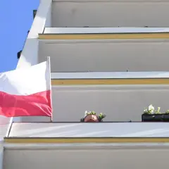 Biało-czerwona bandera polska powiewa na tle błękitnego nieba, obok balkonów z kwiatami.