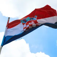 Flaga czerwono biało niebieska z herbem Chorwacji powiewa na wietrze na tle błękitnego nieba z chmurami.