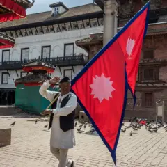 Mężczyzna macha flagą Nepalu na placu pełnym gołębi. W tle tradycyjna architektura.