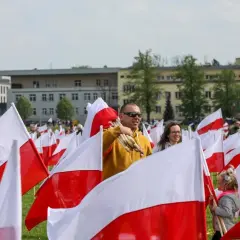 Ludzie z biało-czerwonymi flagami, wśród nich mężczyzna z pęczkiem flag. W tle budynki i drzewa. Flaga Torunia dumnie powiewa.