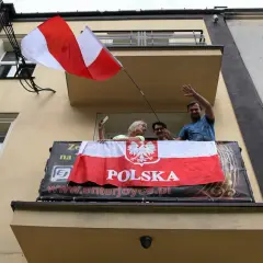 Ludzie na balkonie machają, trzymając polską flagę na maszcie.
