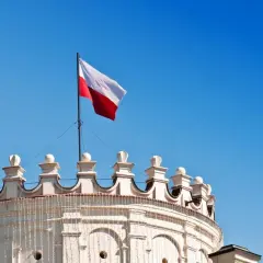 Biało-czerwona flaga Polski powiewa na maszcie nad zabytkowym budynkiem. Wymiary flagi polski są standardowe, a jej kolory symbolizują dumę narodową.