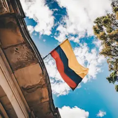 Flaga Kolumbii powiewa na tle błękitnego nieba z chmurami, obok zabytkowego budynku i drzew.