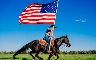 Jeździec w kapeluszu galopuje na czarnym koniu, niosąc pierwszą flagę USA na tle błękitnego nieba.