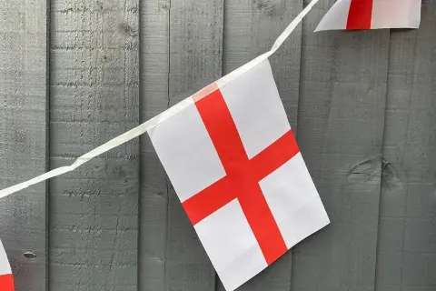 Girlanda z flagami Anglii wisi na szarej, drewnianej desce. Białe tło z czerwonym krzyżem.