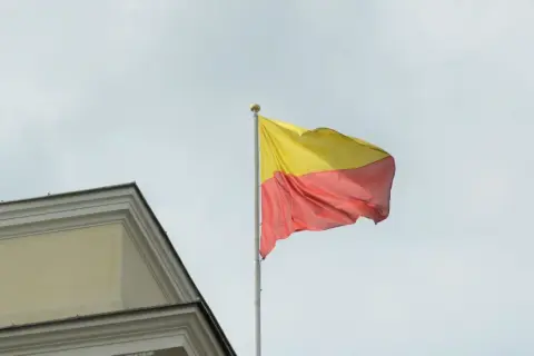 Flaga Warszawy powiewa na maszcie obok zabytkowego budynku.