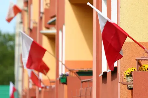Kilka biało-czerwonych flag państwowych powiewa na tle pomarańczowo-żółtego budynku.