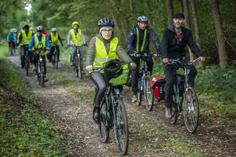 Grupa rowerzystów w kamizelkach odblaskowych jedzie leśną ścieżką. Jedna z pań ma na rowerze zieloną flagę.