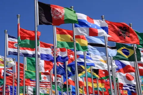 Wiele flag narodowych powiewa na wietrze. Wśród nich flaga biało niebieska z gwiazdami.
