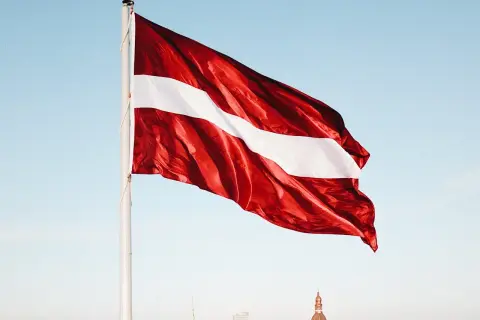 Flaga Łotwy: ciemnoczerwona z białą wstęgą pośrodku, na tle błękitnego nieba i miasta.