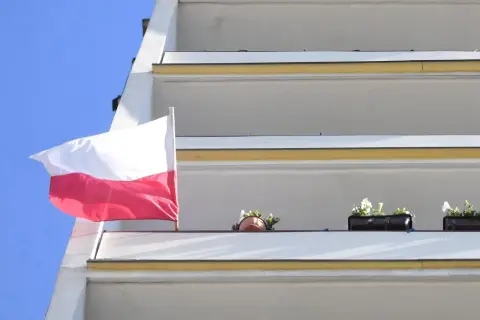 Biało-czerwona bandera polska powiewa na tle błękitnego nieba, obok balkonów z kwiatami.