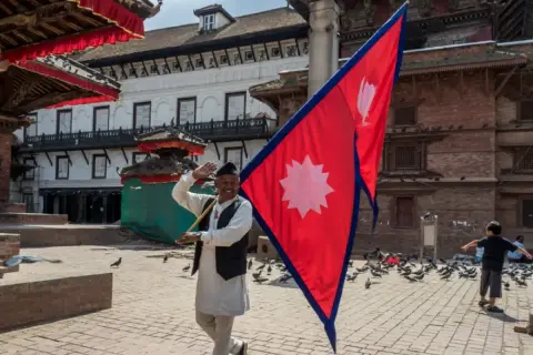Mężczyzna macha flagą Nepalu na placu pełnym gołębi. W tle tradycyjna architektura.