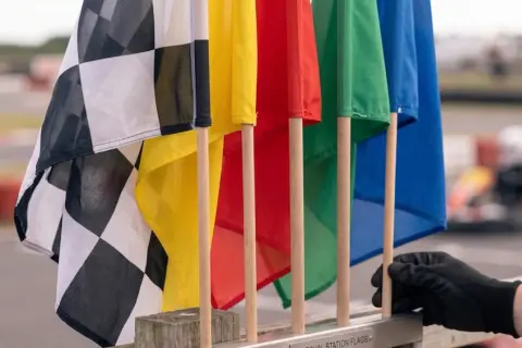 Flaga wyścigowa w kratkę obok kolorowych flag: żółtej, czerwonej, zielonej i niebieskiej. W tle widać tor wyścigowy i gokarta.
