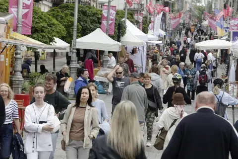 Tłum ludzi spaceruje ulicą z kramami i namiotami, ozdobioną flagami.