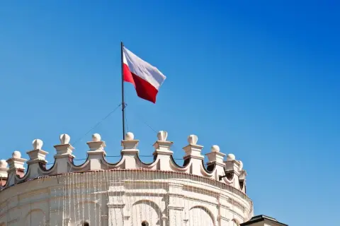 Biało-czerwona flaga Polski powiewa na maszcie nad zabytkowym budynkiem. Wymiary flagi polski są standardowe, a jej kolory symbolizują dumę narodową.