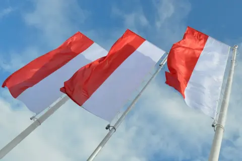 Trzy biało-czerwone flagi powiewające na tle błękitnego nieba z chmurami.