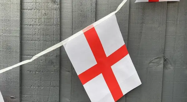 Girlanda z flagami Anglii wisi na szarej, drewnianej desce. Białe tło z czerwonym krzyżem.