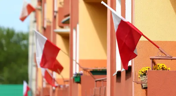 Kilka biało-czerwonych flag państwowych powiewa na tle pomarańczowo-żółtego budynku.