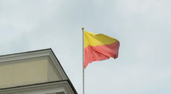 Flaga żółto czerwona powiewa na maszcie obok zabytkowego budynku.