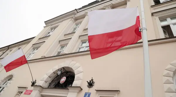 Flaga Polski pionowo powiewa przed budynkiem. Biało-czerwone barwy dumne na tle jasnej fasady.