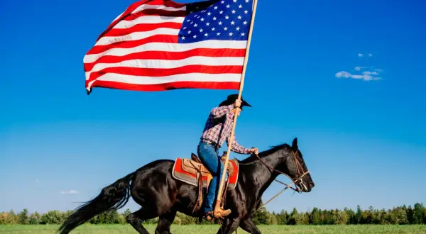 Jeździec w kapeluszu galopuje na czarnym koniu, niosąc pierwszą flagę USA na tle błękitnego nieba.