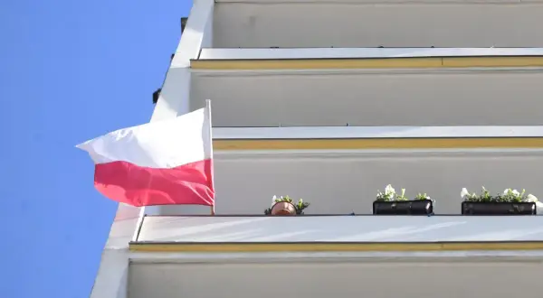 Biało-czerwona bandera polska powiewa na tle błękitnego nieba, obok balkonów z kwiatami.
