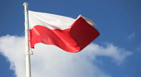 Biało-czerwona flaga Polski powiewa na maszcie na tle błękitnego nieba z chmurami. To symbol ważnych dat, kiedy wywieszamy flagę.