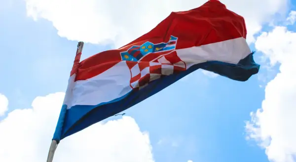 Flaga czerwono biało niebieska z herbem Chorwacji powiewa na wietrze na tle błękitnego nieba z chmurami.