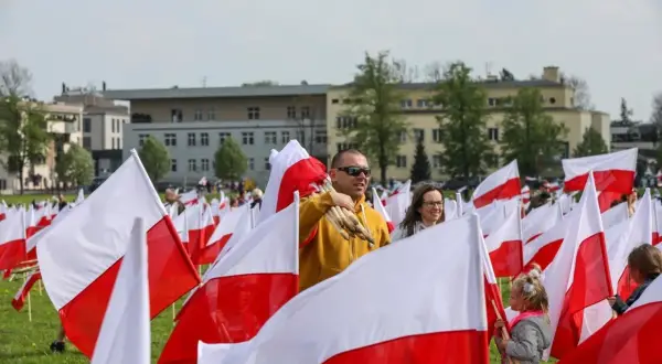 Ludzie z biało-czerwonymi flagami, wśród nich mężczyzna z pęczkiem flag. W tle budynki i drzewa. Flaga Torunia dumnie powiewa.