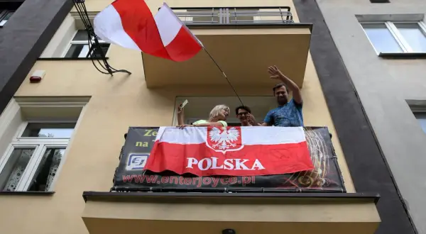 Ludzie na balkonie machają, trzymając polską flagę na maszcie.