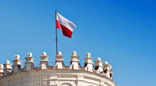 Biało-czerwona flaga Polski powiewa na maszcie nad zabytkowym budynkiem. Wymiary flagi polski są standardowe, a jej kolory symbolizują dumę narodową.