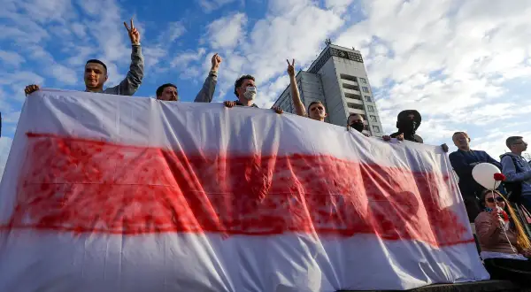 Ludzie trzymają biało-czerwoną flagę, symbol wolności Białorusi. Niebo z chmurami nad budynkiem.