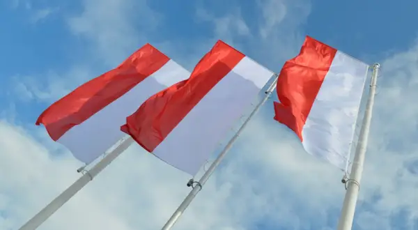 Trzy biało-czerwone flagi powiewające na tle błękitnego nieba z chmurami.