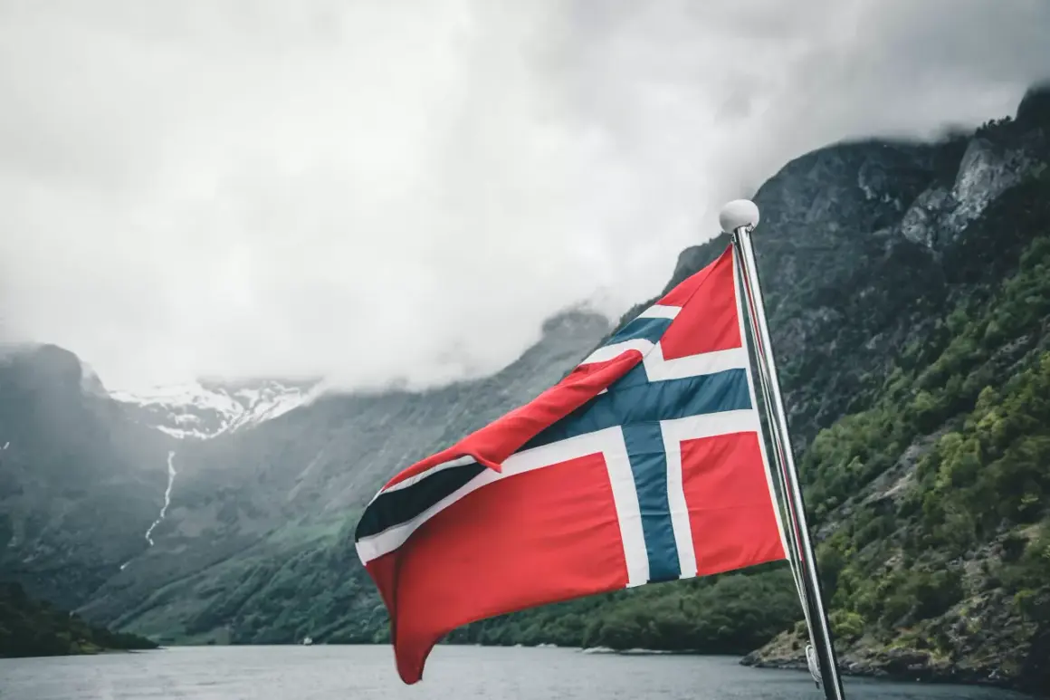 Flaga Norwegii powiewa na maszcie na tle malowniczego fiordu.