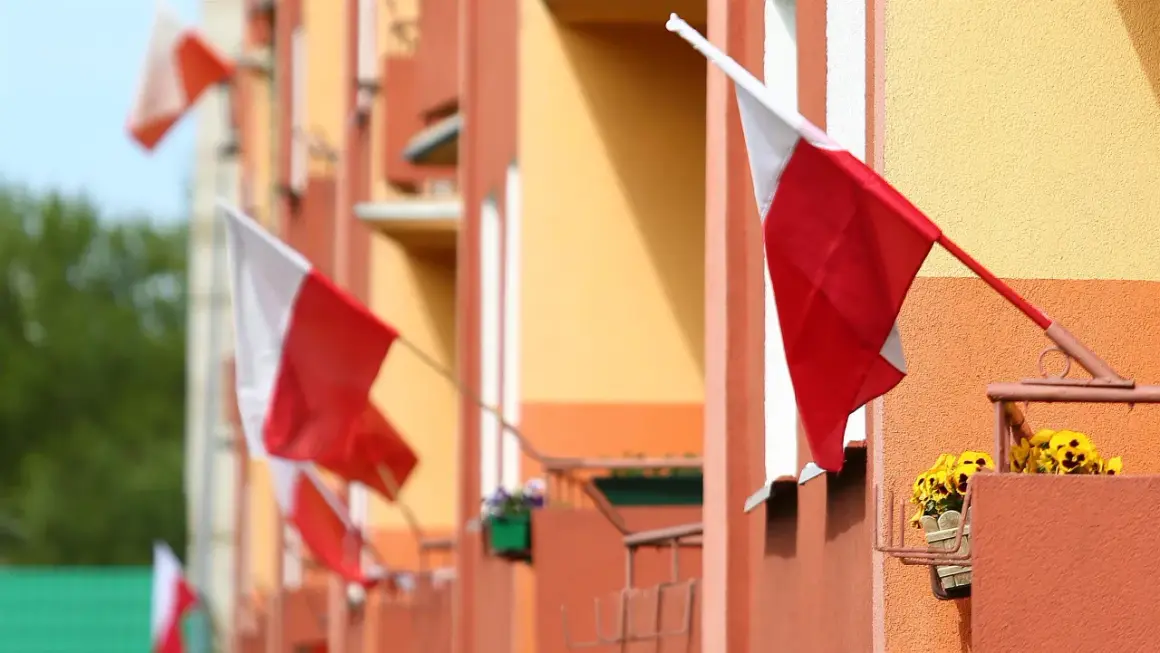 Kilka biało-czerwonych flag państwowych powiewa na tle pomarańczowo-żółtego budynku.