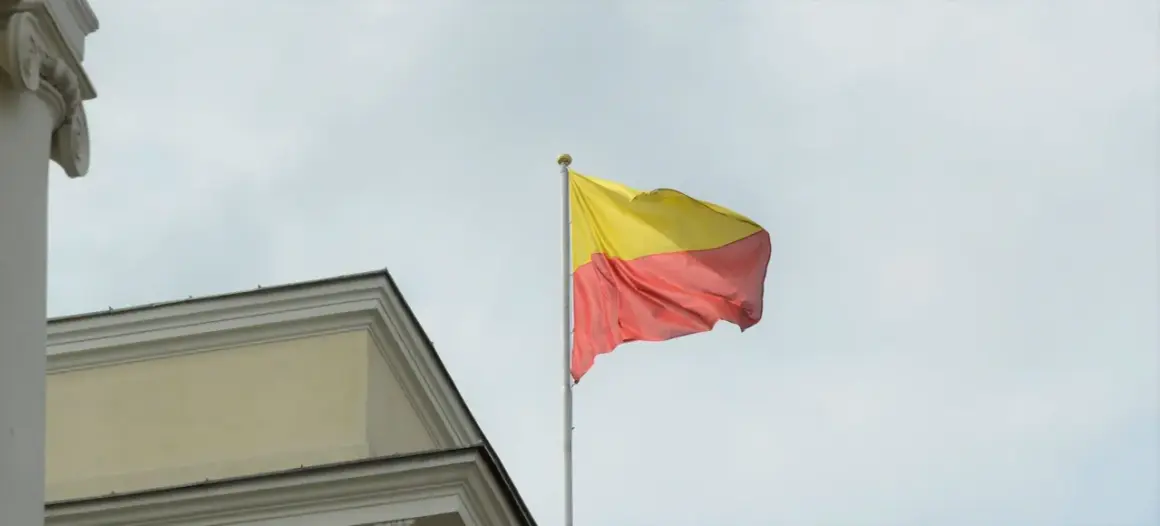 Flaga żółto czerwona powiewa na maszcie obok zabytkowego budynku.