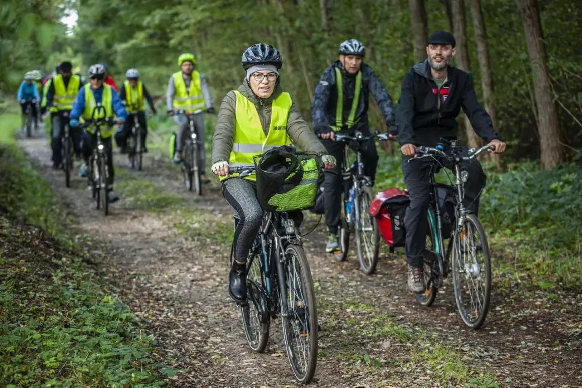 Grupa rowerzystów w kamizelkach odblaskowych jedzie leśną ścieżką. Jedna z pań ma na rowerze zieloną flagę.