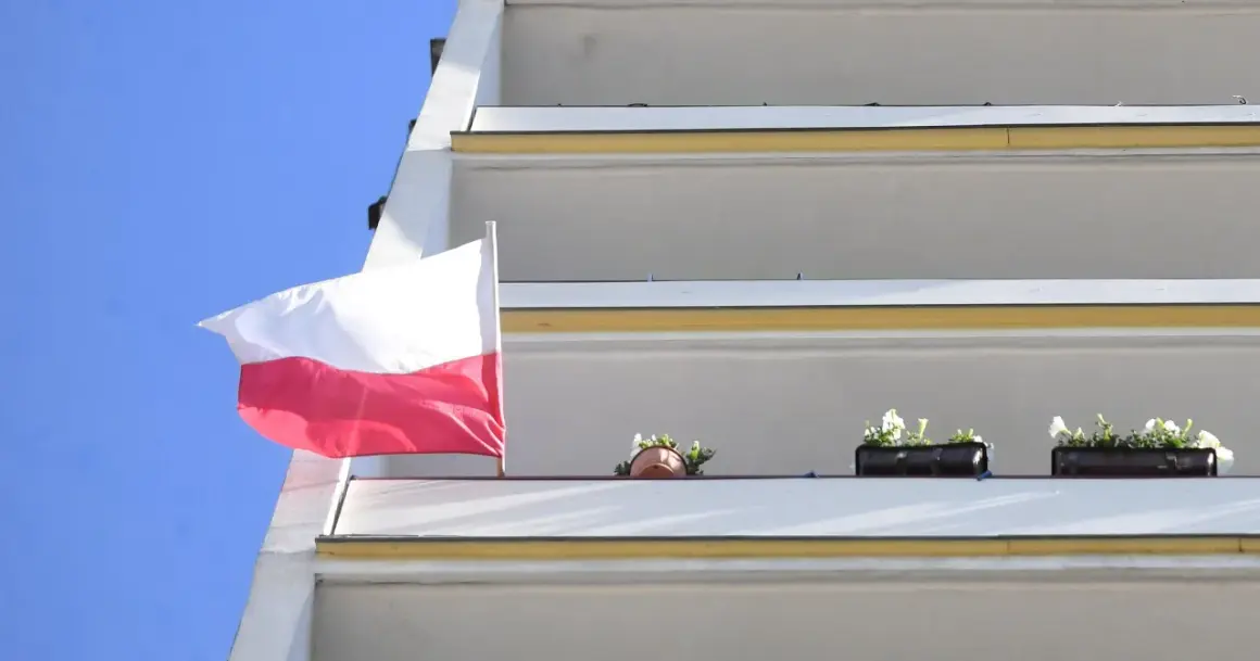 Biało-czerwona bandera polska powiewa na tle błękitnego nieba, obok balkonów z kwiatami.