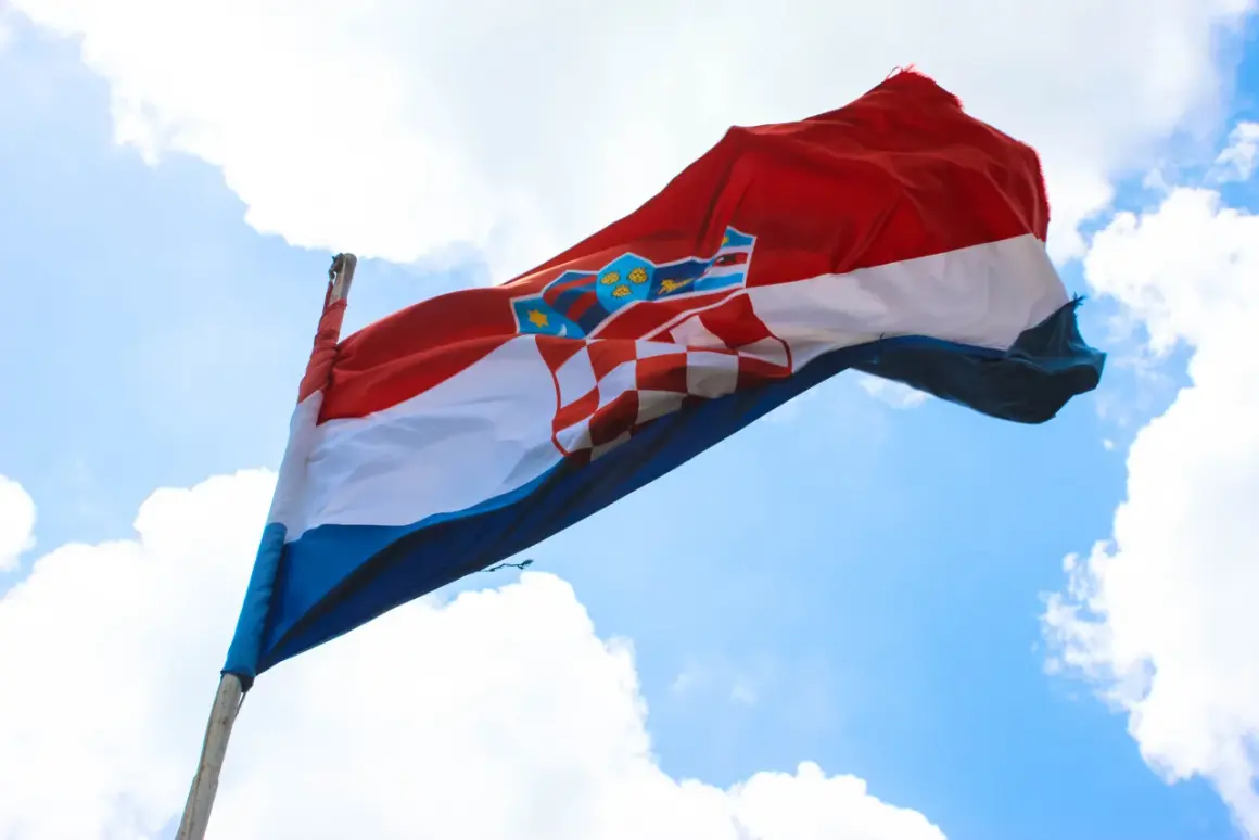 Flaga czerwono biało niebieska z herbem Chorwacji powiewa na wietrze na tle błękitnego nieba z chmurami.