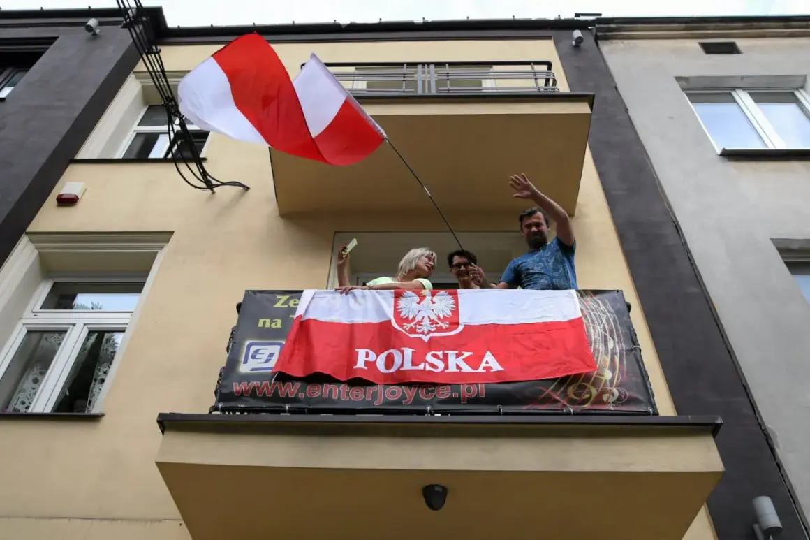 Ludzie na balkonie machają, trzymając polską flagę na maszcie.