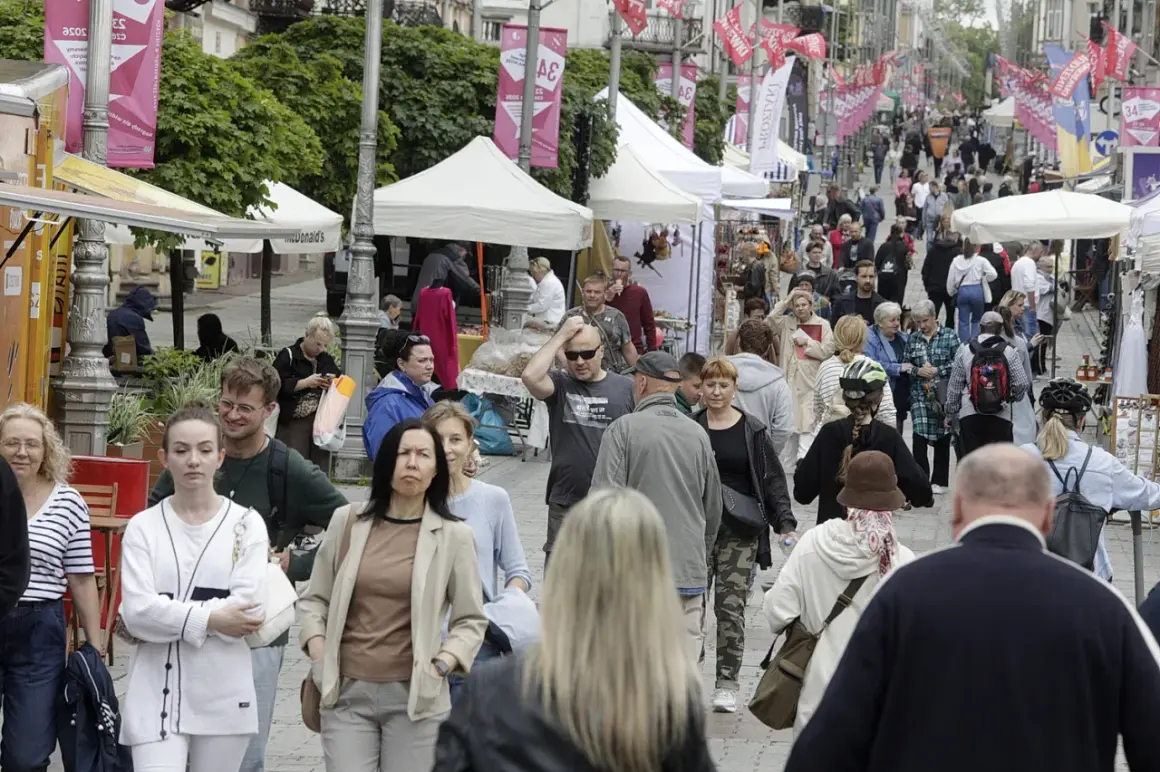 Tłum ludzi spaceruje ulicą z kramami i namiotami, ozdobioną flagami.