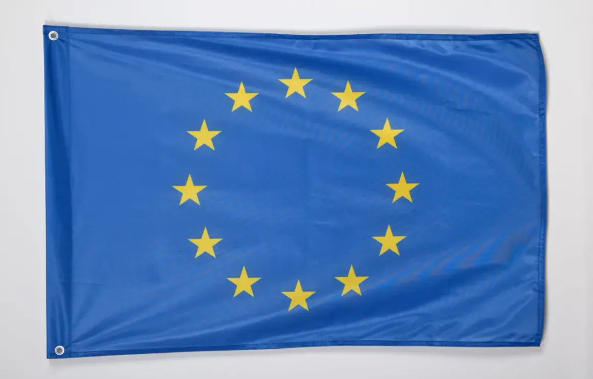 Niebieska flaga z dwunastoma złotymi gwiazdami ułożonymi w okrąg. To godło Unii Europejskiej, symbol jedności i solidarności.