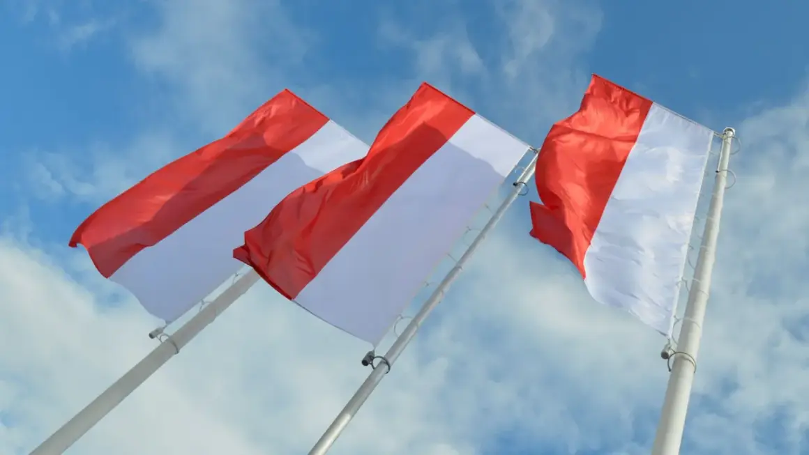 Trzy biało-czerwone flagi powiewające na tle błękitnego nieba z chmurami.