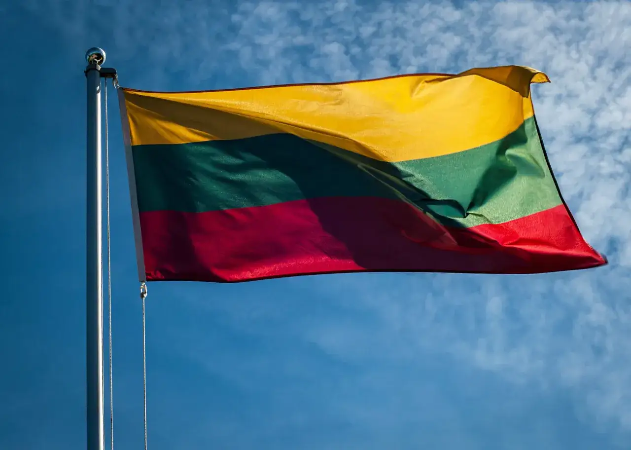 Flaga Litwy powiewa na maszcie na tle błękitnego nieba z chmurami.